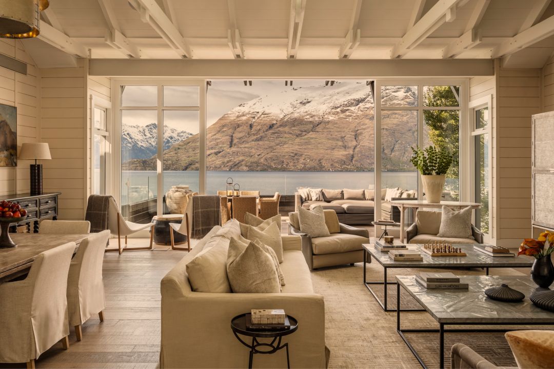 Spacious living and dining area in a luxury four-bedroom villa with neutral furnishings and doors leading out to a large private deck that overlooks the lake and snow-capped mountains.